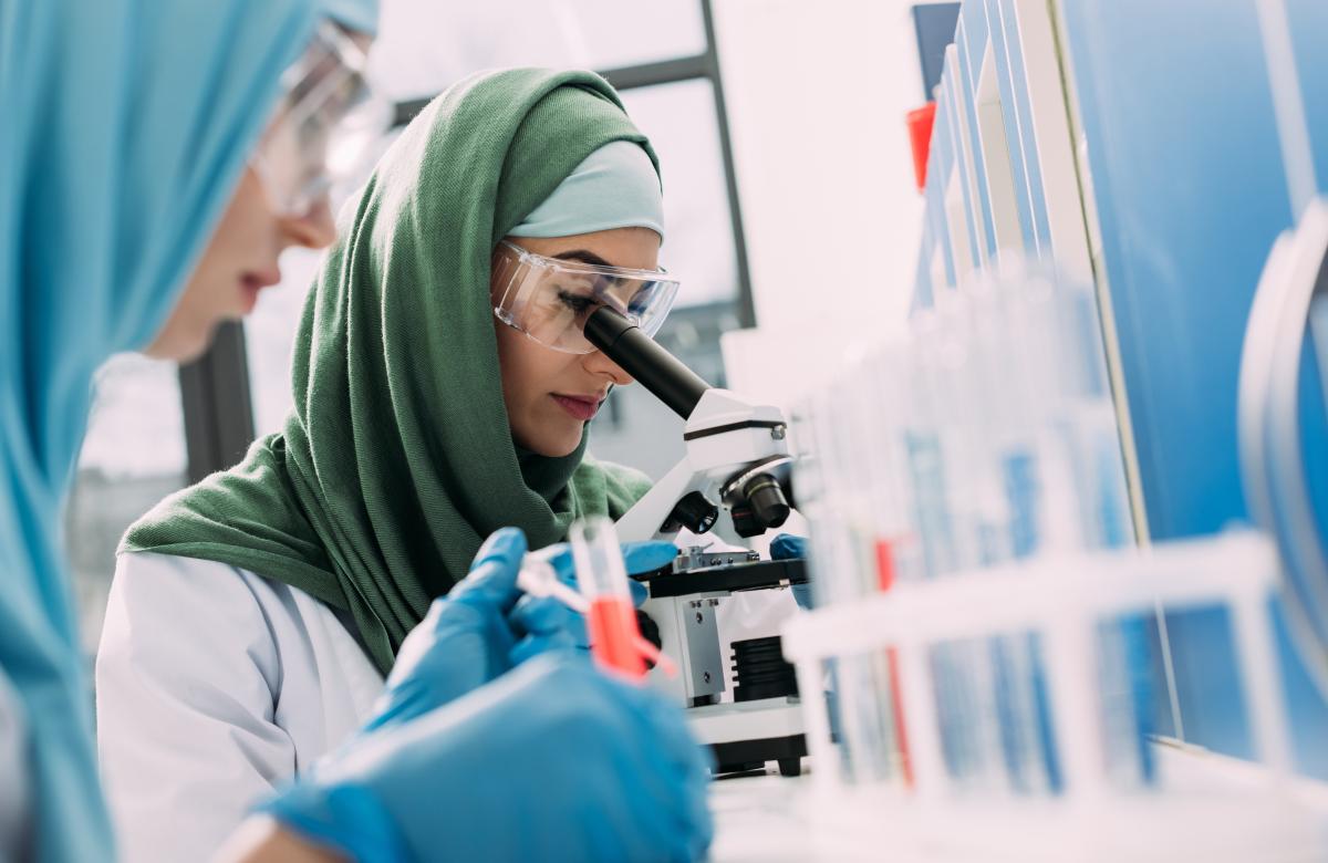 A photo of two labworkers, one looking into a microscope and one looking at test tubes.